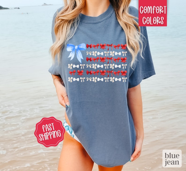 May include: Blue jean colored t-shirt with a watercolor design of a flag made of bows. The flag design features a large blue bow in the upper left corner and rows of red and white bows. The words "Comfort Colors" are in the upper right corner.