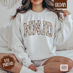 May include: A light gray sweatshirt with the letters "WWJD" in a camouflage pattern. The sweatshirt is a crew neck style and is made of a soft material. The brand name "Gildan" is visible in the upper right corner.