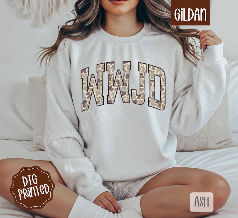 May include: A light gray sweatshirt with the letters "WWJD" in a camouflage pattern. The sweatshirt is a crew neck style and is made of a soft material. The brand name "Gildan" is visible in the upper right corner.