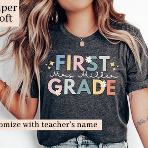 Op de afbeelding: Een grijs T-shirt met de tekst "FIRST" in blauw, "Mrs. Miller" in roze en "GRADE" in geel en groen. De tekst is versierd met witte madeliefjes en gouden sterren. Het shirt is te personaliseren met de naam van een leraar.