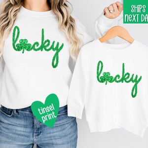 May include: White sweatshirt with green glitter "lucky" text and a shamrock.  The sweatshirt is paired with a white t-shirt with the same design.  The text is in a cursive font.