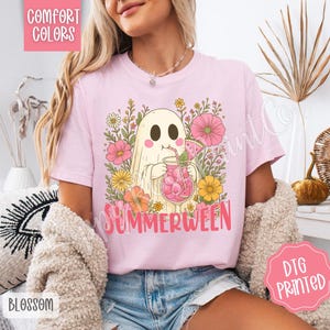May include: Light pink t-shirt with a graphic of a ghost drinking a pink beverage, surrounded by flowers. The word "SUMMERWEEN" is printed in pink. The shirt has the words "COMFORT COLORS" and "DTG PRINTED" printed on it.