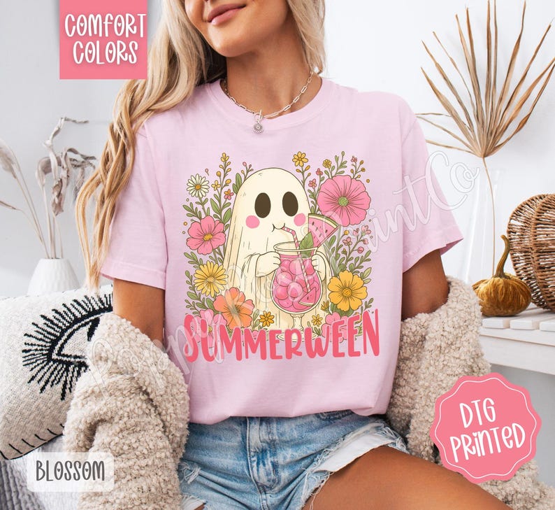 May include: Light pink t-shirt with a graphic of a ghost drinking a pink beverage, surrounded by flowers. The word "SUMMERWEEN" is printed in pink. The shirt has the words "COMFORT COLORS" and "DTG PRINTED" printed on it.