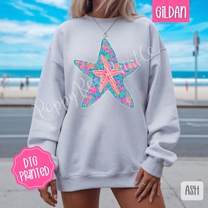 May include: A light gray sweatshirt with a floral starfish design. The sweatshirt is made by Gildan and is DTG printed. The text "ASH" is visible in the lower right corner of the image.