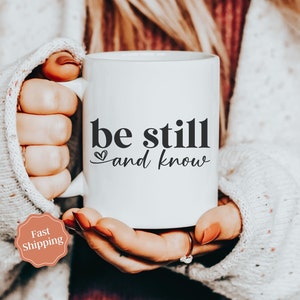 May include: White ceramic mug with black text that reads "be still and know" with a small heart symbol. The mug is being held by a person wearing a white sweater.