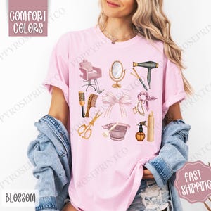 May include: A pink t-shirt with a graphic design of hair salon tools and equipment, including a hair dryer, scissors, a hairbrush, a hair dye bowl, a hairspray bottle, a mirror, a hair tie, and a salon chair. The text "COMFORT COLORS" is at the top of the shirt.