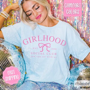 May include: A light blue t-shirt with the text "GIRLHOOD EST 1988 SOCIAL CLUB you can sit with us" in pink lettering. The shirt features a pink bow graphic.