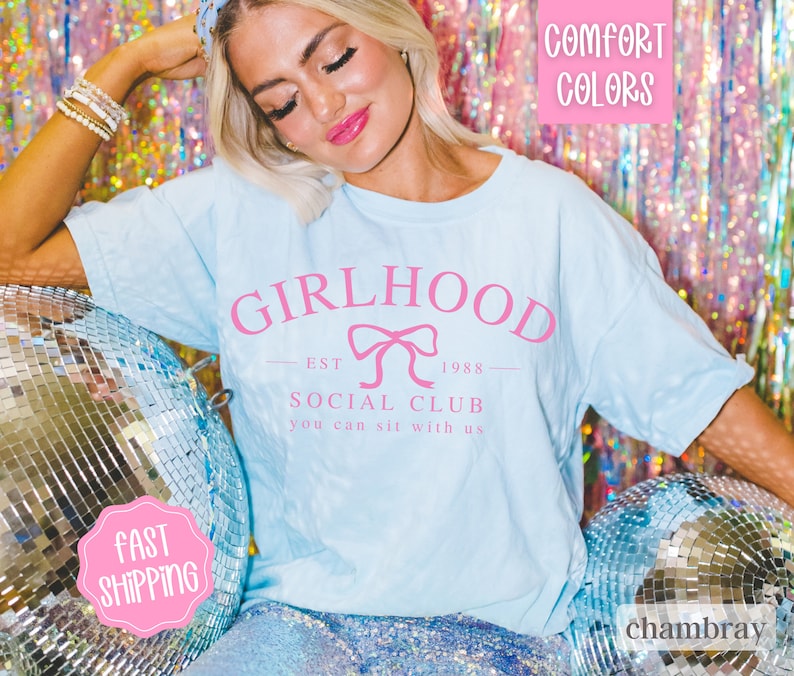 May include: A light blue t-shirt with the text "GIRLHOOD EST 1988 SOCIAL CLUB you can sit with us" in pink lettering. The shirt features a pink bow graphic.