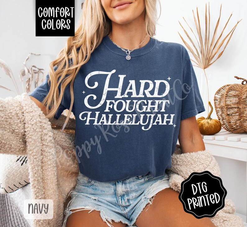 May include: Navy blue t-shirt with the words "Hard Fought Hallelujah" in white script. The shirt is a Comfort Colors brand and has a DTG printed design. The shirt is paired with denim shorts.