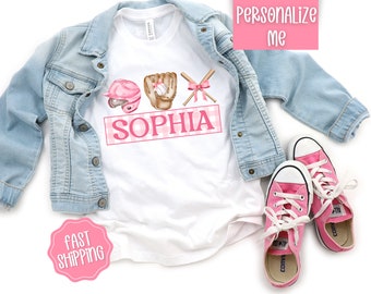 May include: White t-shirt with a pink and white gingham background. The shirt features a pink baseball helmet, glove, bats, and a bow. The name "Sophia" is written in pink letters on the shirt.