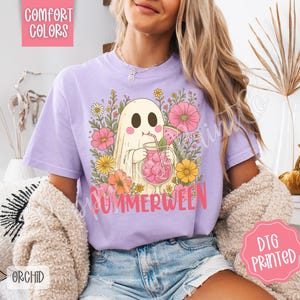 May include: A light purple t-shirt with a graphic of a ghost drinking a pink beverage with watermelon slices, surrounded by flowers. The word "SUMMERWEEN" is printed in pink. The shirt also has the words "COMFORT COLORS" and "ORCHID" printed on it.