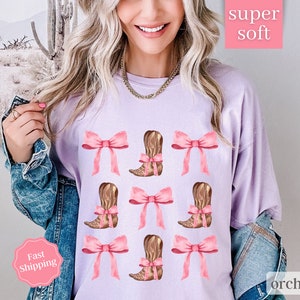 May include: A lavender t-shirt with a pattern of pink bows and brown cowboy boots. The text "super soft" and "orchid" are printed on the shirt.