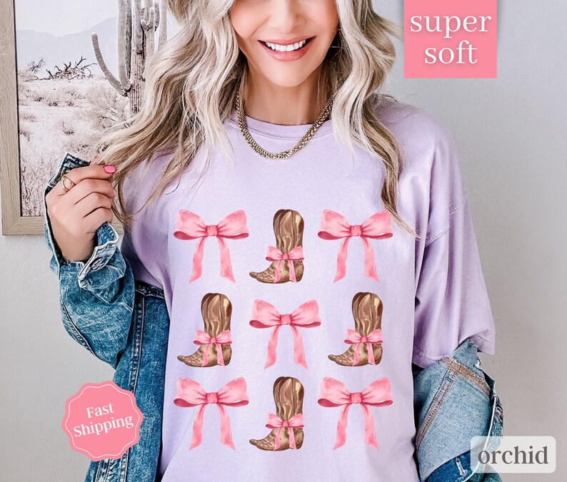 May include: A lavender t-shirt with a pattern of pink bows and brown cowboy boots. The text "super soft" and "orchid" are printed on the shirt.