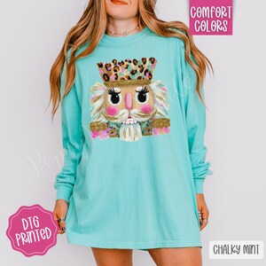 May include: A long-sleeved, turquoise shirt featuring a colorful nutcracker graphic. The nutcracker has a leopard print crown, pink cheeks, and a white beard. The shirt also has the words "Comfort Colors" and "Chalky Mint" printed on it.
