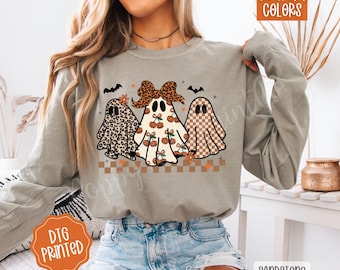 May include: Sandstone long-sleeve shirt featuring three cartoon ghosts with different patterns and designs. The ghosts have a leopard print, cherry design, and checkered pattern. The shirt also has the words "Comfort Colors" and "DTG Printed".