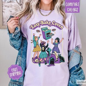 May include: A light purple t-shirt featuring a graphic print with the text Easy-Bake Coven. The illustration depicts three figures, a stove, a black cat, a tombstone with RIP, and skulls. The shirt is worn under a blue denim jacket.