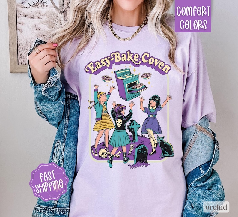 May include: A light purple t-shirt featuring a graphic print with the text Easy-Bake Coven. The illustration depicts three figures, a stove, a black cat, a tombstone with RIP, and skulls. The shirt is worn under a blue denim jacket.
