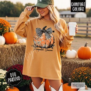 May include: Mustard-colored long-sleeve shirt with a pumpkin graphic. The pumpkin design features a black and white checkered bow and floral accents. The text "Pumpkin Season" is printed below the pumpkin. The shirt is labeled "DTG Printed" and "Comfort Colors".