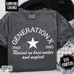 May include: Dark grey long-sleeve shirt with the text "GENERATION X" in a semi-circle, a star, and the years 1965 and 1980. The phrase "Raised on hose water and neglect" is below. The shirt is labeled "Comfort Colors" and "DTG Printed".
