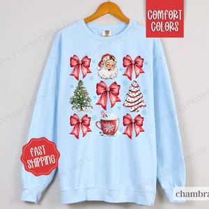 May include: A light blue sweatshirt with a Christmas theme featuring a Santa Claus, Christmas trees, bows, and a cup of hot chocolate. The text "Comfort Colors" is in the top right corner of the image.