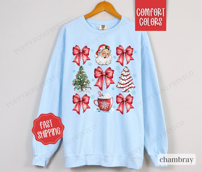 May include: A light blue sweatshirt with a Christmas theme featuring a Santa Claus, Christmas trees, bows, and a cup of hot chocolate. The text "Comfort Colors" is in the top right corner of the image.