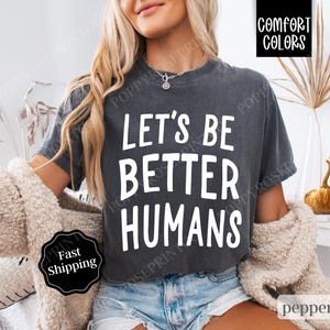 May include: A dark gray cropped t-shirt with the text "Let's Be Better Humans" printed in white. The shirt is being worn by a person.