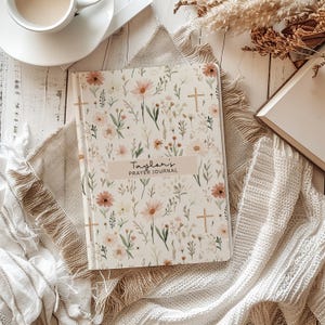 May include: A floral-patterned prayer journal with the text "Taylor's Prayer Journal" on the cover. The cover features a cream background with pink and white flowers, green leaves, and gold crosses. The journal is open on a beige cloth.
