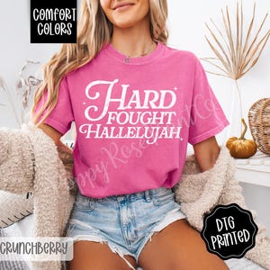 May include: A pink Comfort Colors t-shirt with the words "Hard Fought Hallelujah" in white script. The shirt is paired with denim shorts and a cream-colored cardigan. The image also includes a black and white "DTG Printed" sticker.