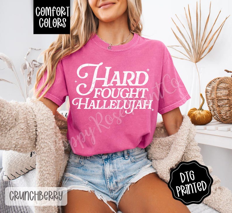 May include: A pink Comfort Colors t-shirt with the words "Hard Fought Hallelujah" in white script. The shirt is paired with denim shorts and a cream-colored cardigan. The image also includes a black and white "DTG Printed" sticker.
