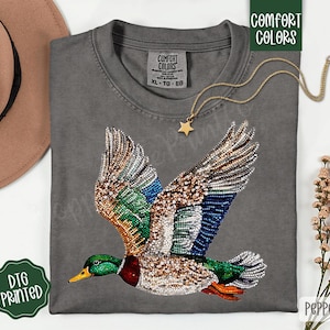 May include: Gray t-shirt with a detailed, bejeweled duck design in flight. The duck features green, blue, brown, and white accents. The shirt has a crew neck and a tag that says "Comfort Colors". A gold necklace with a star pendant is also visible.