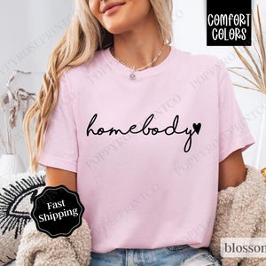 May include: Pink t-shirt with the text "homebody" in black script with a small heart next to it.