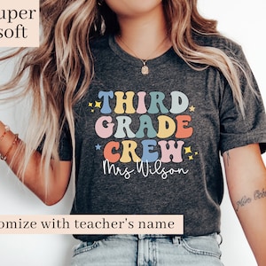 May include: A dark grey t-shirt with the text "THIRD GRADE CREW" in colourful, retro-style letters. The text "Mrs. Wilson" is written below the main text. The shirt is being worn by a person with long blonde hair.