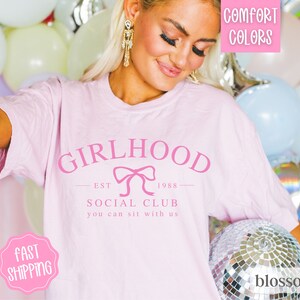 May include: Pink t-shirt with the text "Girlhood Est 1988 Social Club You can sit with us" and a pink bow graphic.