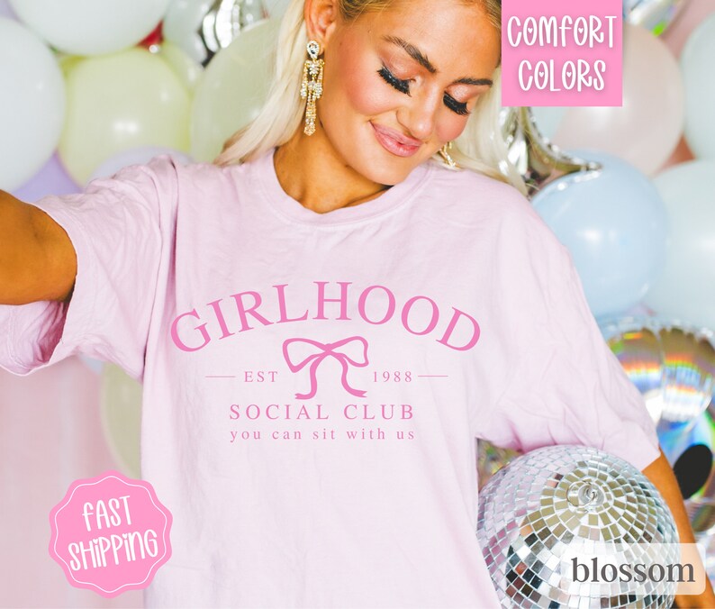 May include: Pink t-shirt with the text "Girlhood Est 1988 Social Club You can sit with us" and a pink bow graphic.