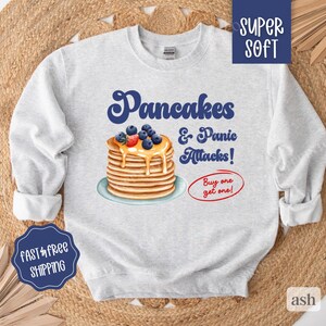 May include: A gray sweatshirt with a graphic of a stack of pancakes with blueberries and syrup, and the text "Pancakes & Panic Attacks! Buy one get one!"