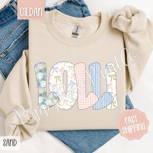 May include: A beige Gildan sweatshirt with the name "Loli" spelled out in colorful patchwork letters. The letters are made of different fabrics with floral, gingham, and striped patterns.