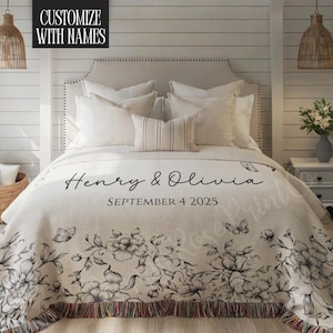 May include: A cream-colored throw blanket with a floral design and the names "Henry & Olivia" and the date "September 4 2025" printed on it. The blanket has a fringed edge and is on a bed with white pillows.