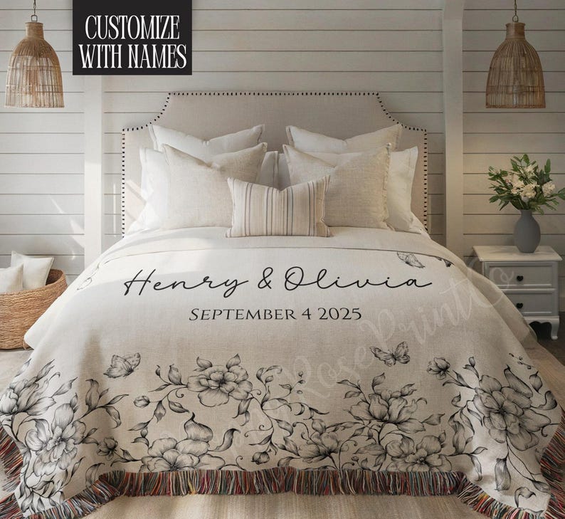 May include: A cream-colored throw blanket with a floral design and the names "Henry & Olivia" and the date "September 4 2025" printed on it. The blanket has a fringed edge and is on a bed with white pillows.