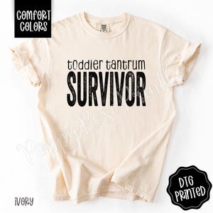 May include: Ivory-colored t-shirt with the words "toddler tantrum SURVIVOR" in black, distressed font. The shirt has rolled-up sleeves and is labeled "Comfort Colors" and "DTG Printed".