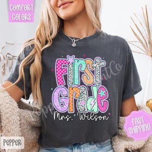May include: Dark grey t-shirt featuring a vibrant "First Grade" design. The graphic includes stars, lightning bolts, and the name "Mrs. Wilson". The text is in various patterns and colours, including pink, orange, and teal.