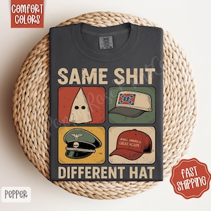 May include: Dark grey t-shirt featuring the text "SAME SHIT DIFFERENT HAT" and illustrations of a KKK hood, a Confederate flag hat, a Nazi hat, and a "Make America Great Again" hat. The shirt is displayed on a woven surface.
