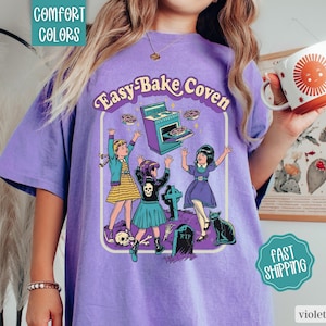 May include: A purple t-shirt featuring a graphic print with the text Easy-Bake Coven. The illustration depicts three children, a stove, a tombstone, a skull, and a black cat. The shirt has a crew neckline and short sleeves, showcasing a vintage-inspired design.