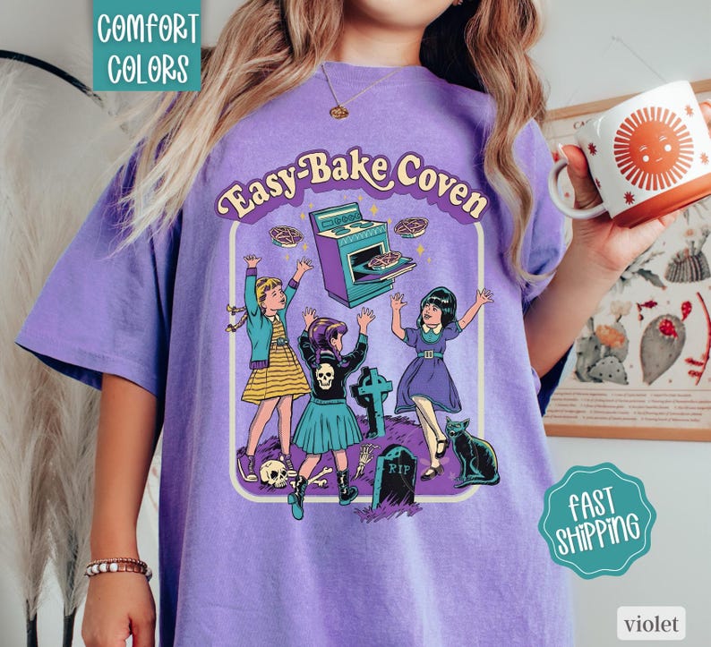 May include: A purple t-shirt featuring a graphic print with the text Easy-Bake Coven. The illustration depicts three children, a stove, a tombstone, a skull, and a black cat. The shirt has a crew neckline and short sleeves, showcasing a vintage-inspired design.