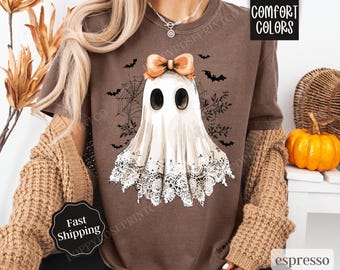 May include: Brown t-shirt with a white ghost wearing a lace trim and an orange bow. The ghost has black eyes and is surrounded by bats and spiderwebs. The text "COMFORT COLORS" is in the top right corner of the image.
