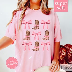 May include: Pink t-shirt with a graphic of brown cowboy boots with pink bows. The text "super soft" is in the top right corner of the image. The text "blossom" is in the bottom right corner of the image.