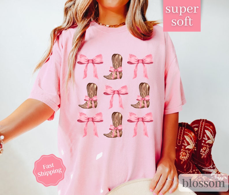 May include: Pink t-shirt with a graphic of brown cowboy boots with pink bows. The text "super soft" is in the top right corner of the image. The text "blossom" is in the bottom right corner of the image.