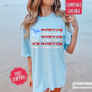 May include: Light blue t-shirt with a watercolor design of an American flag made of bows. The flag design features a large blue bow in the upper left corner and rows of red and white bows. The shirt has the words "Comfort Colors" and "chambray" printed on it.