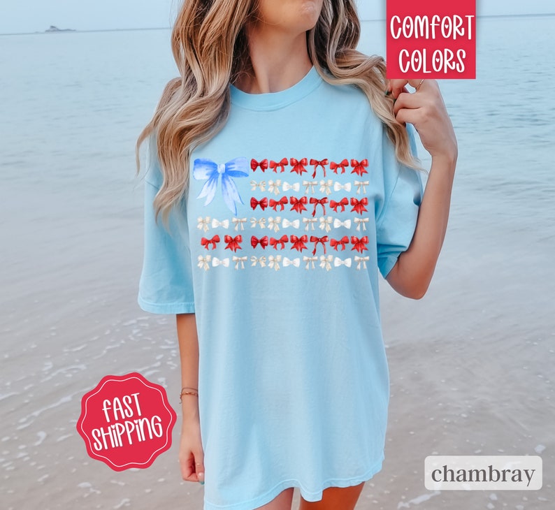 May include: Light blue t-shirt with a watercolor design of an American flag made of bows. The flag design features a large blue bow in the upper left corner and rows of red and white bows. The shirt has the words "Comfort Colors" and "chambray" printed on it.