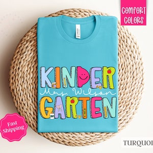 May include: Turquoise blue t-shirt with the text "Kinder" in colorful letters, followed by "Mrs. Wilson" and "Garten" in colorful letters. The letters are decorated with school-themed illustrations.