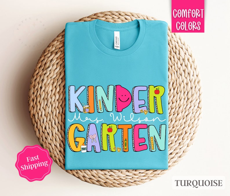 May include: Turquoise blue t-shirt with the text "Kinder" in colorful letters, followed by "Mrs. Wilson" and "Garten" in colorful letters. The letters are decorated with school-themed illustrations.